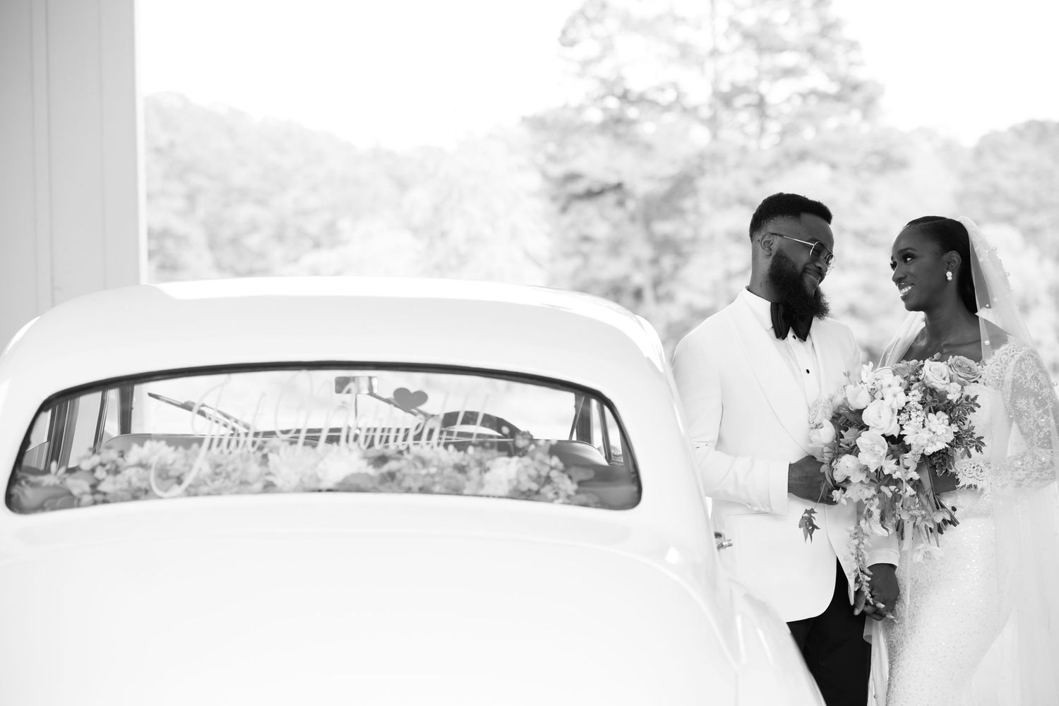 Black and white wedding photo of bride and groom standing beside a vintage car, bride holding a bouquet.