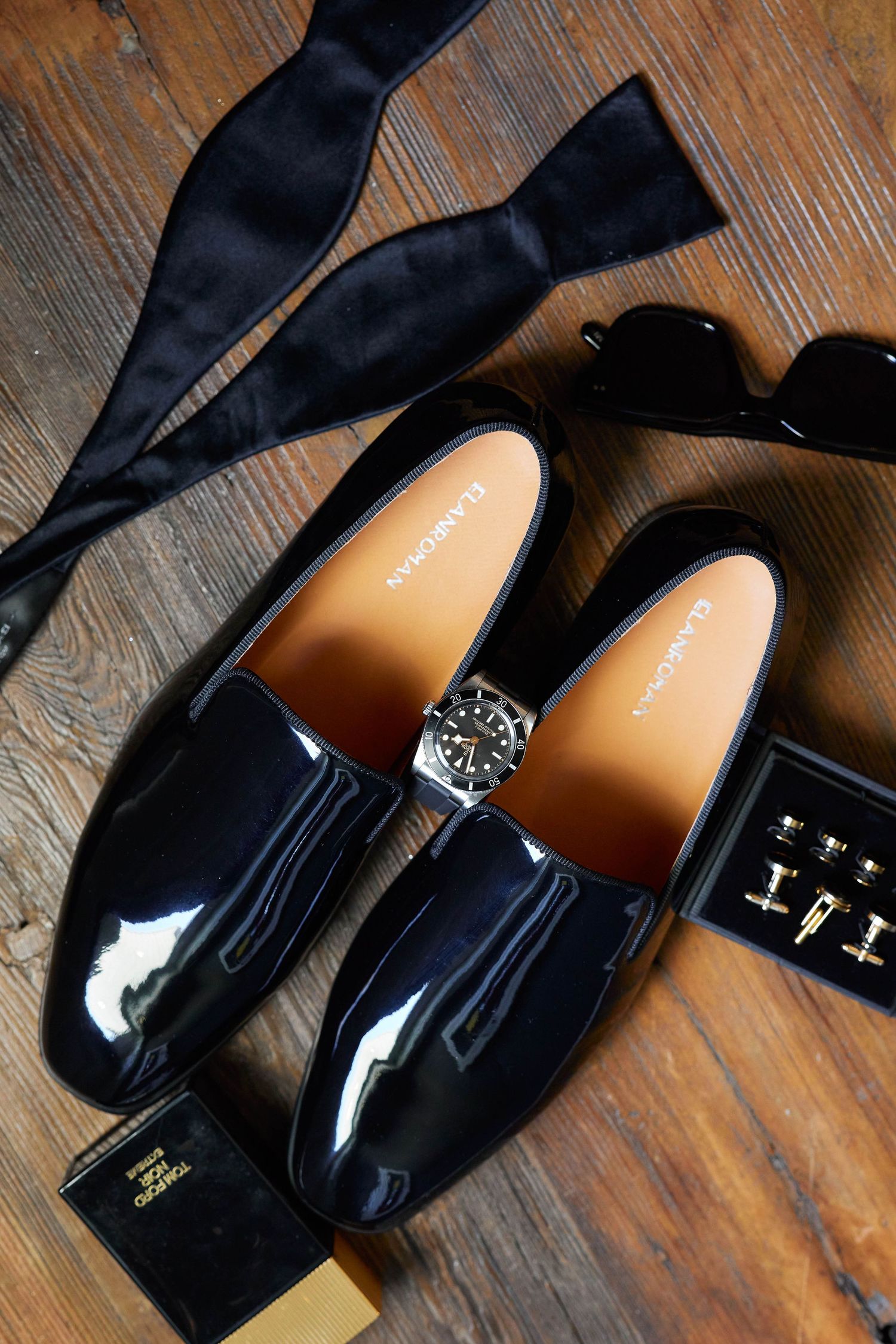 Black patent leather loafers displayed with bow tie, watch, cufflinks, and sunglasses on wooden surface.