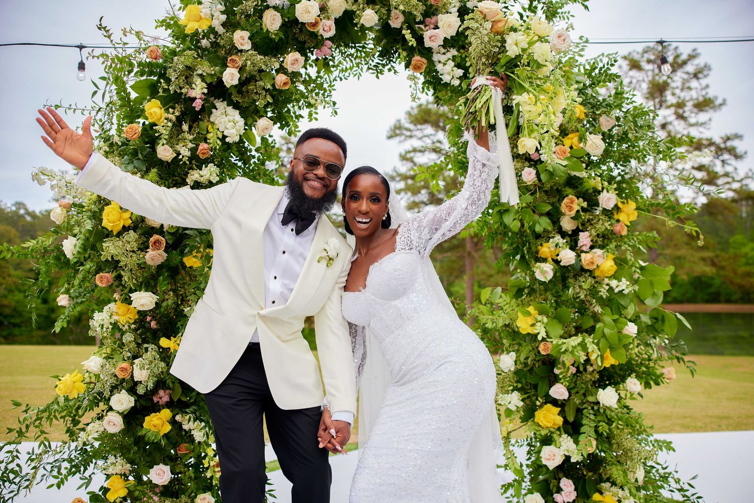 Happy couple in white attire posing under a floral arch with yellow and pink flowers at their outdoor wedding.