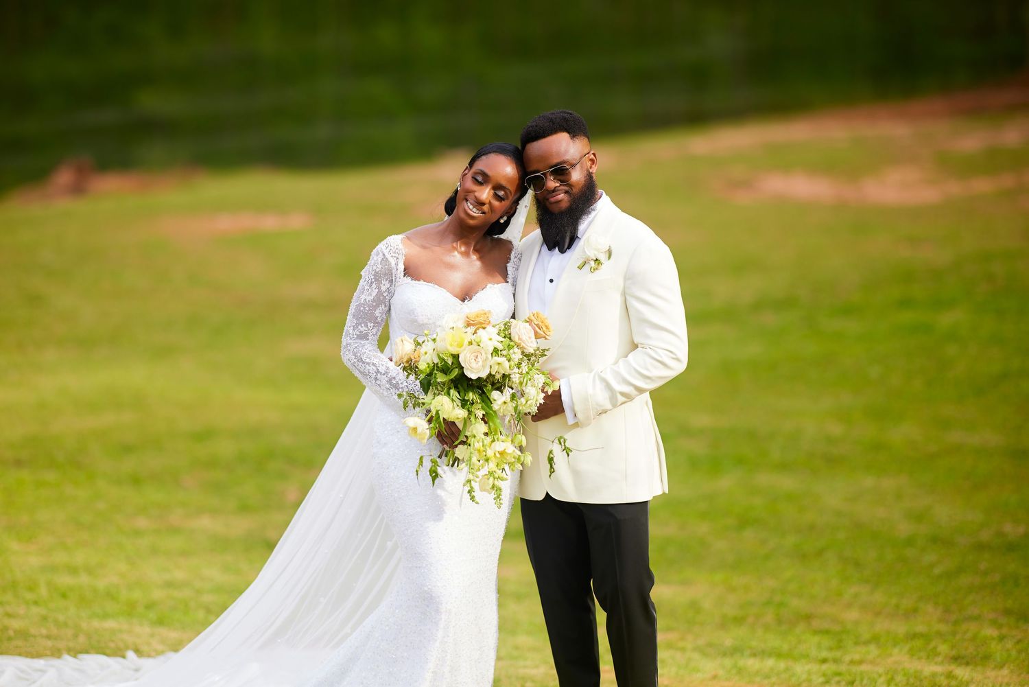 Bride and groom posing outdoors on green grass, bride in white gown holding bouquet, groom in white tuxedo jacket.