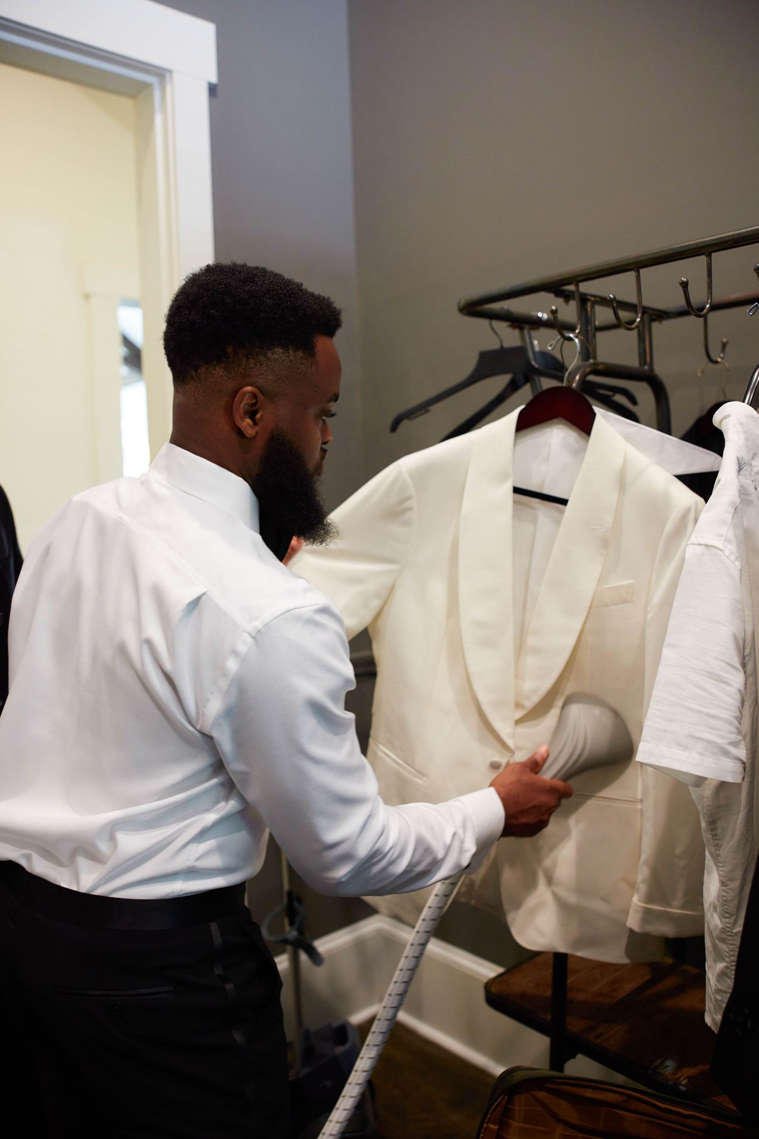A man steams a white tuxedo jacket on a hanger in a dressing room before a formal event.