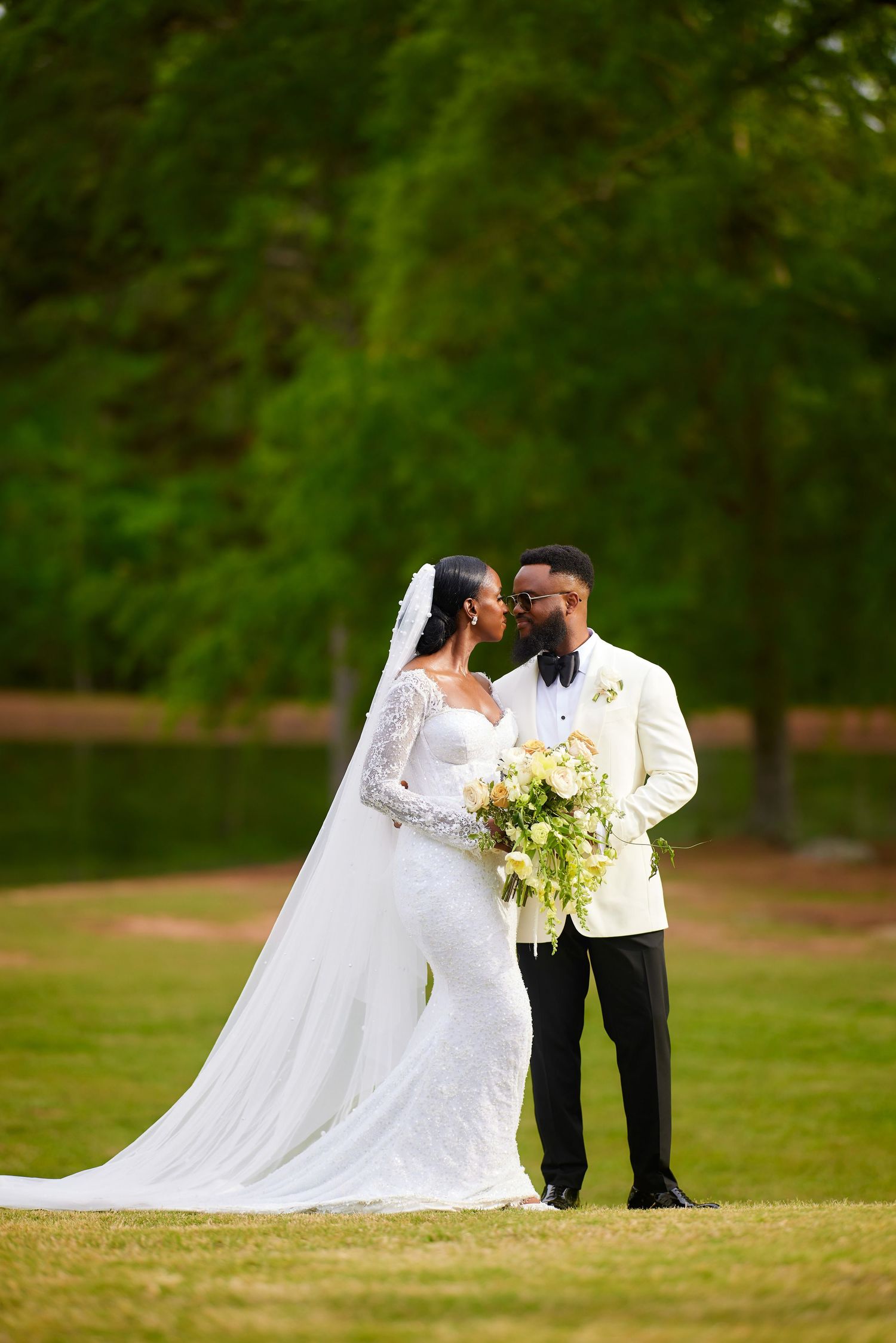 Bride and groom share a romantic moment outdoors, bride in lace gown with veil, groom in white tuxedo jacket.