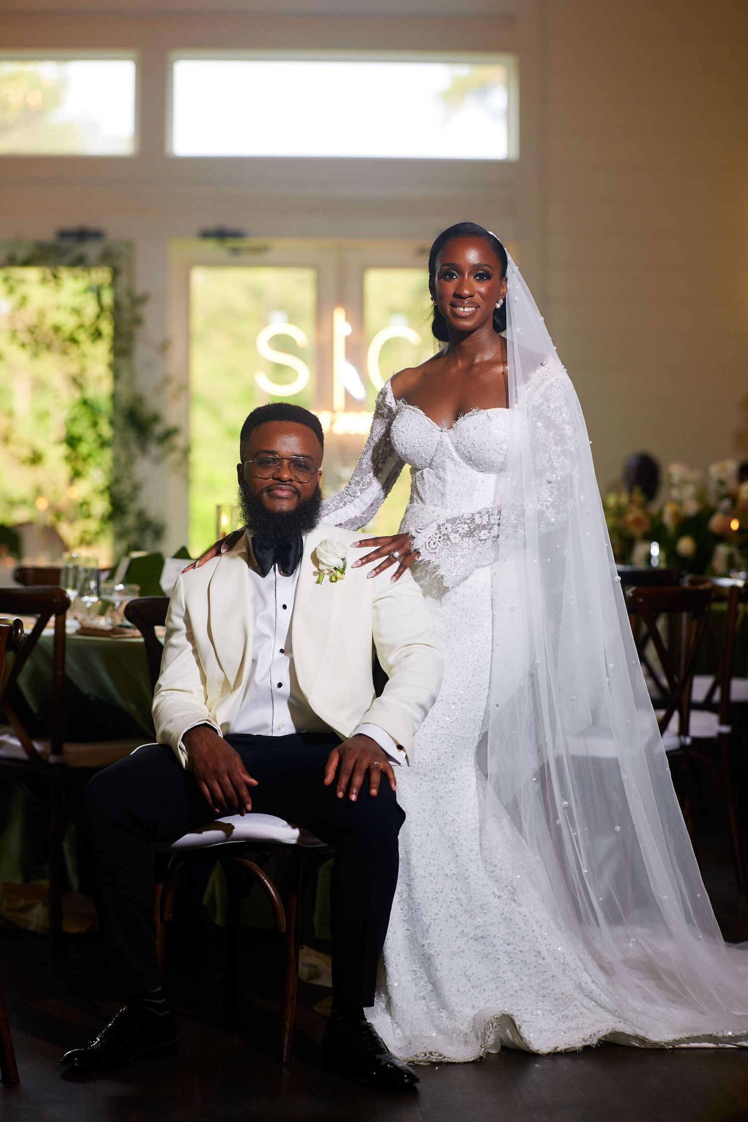 Elegant bride and groom pose at wedding reception, bride in lace gown with veil, groom in white tuxedo jacket.