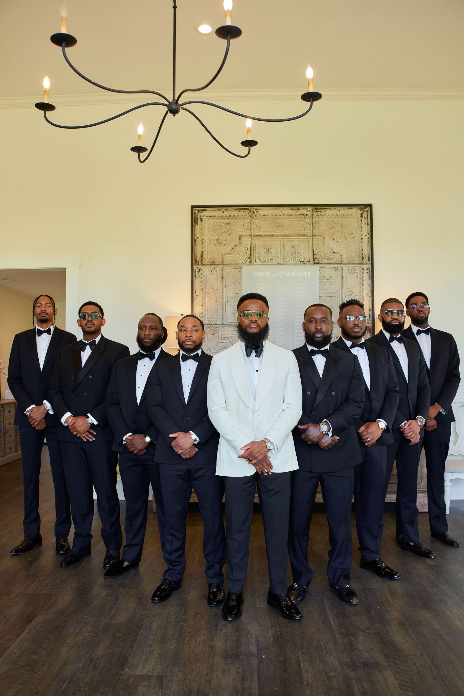 Groom in white tuxedo jacket stands with groomsmen in black tuxedos under chandelier in elegant venue.