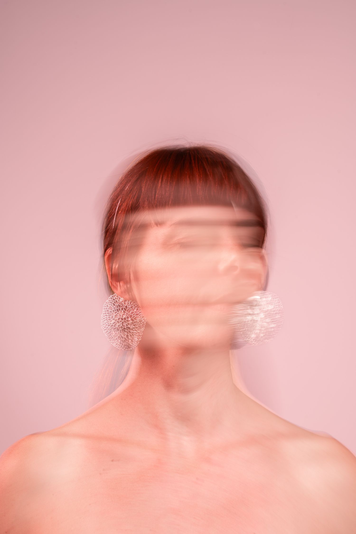 Blurred motion portrait of a redhead woman with bangs wearing crystal earrings against a pink background.