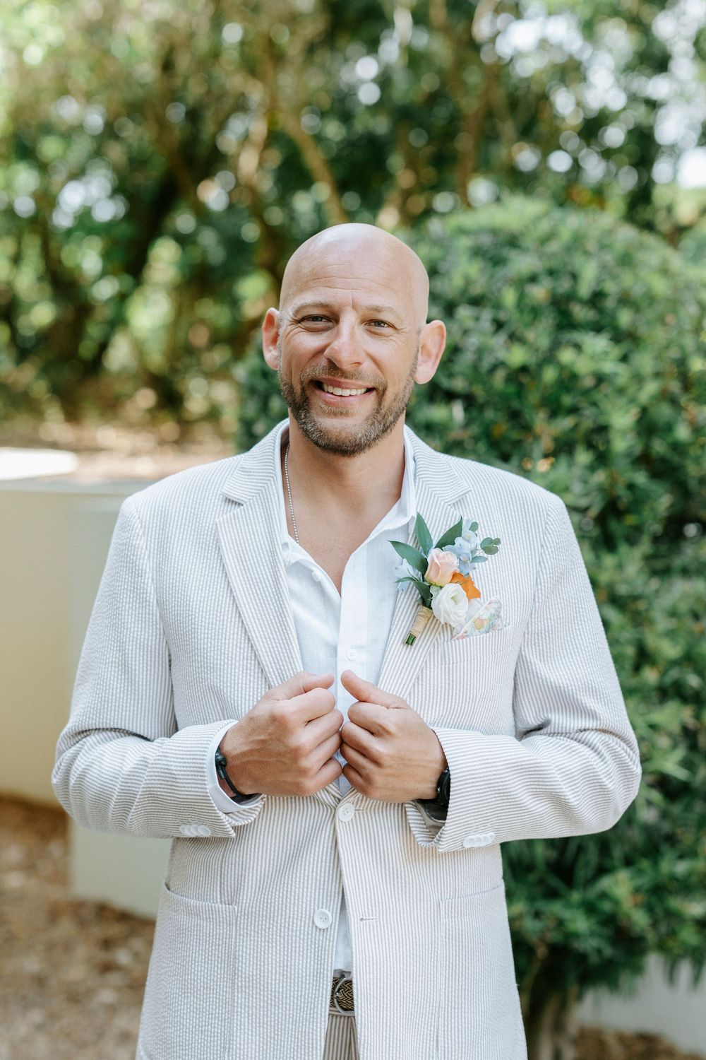 Smiling bald groom in light gray seersucker suit with floral boutonniere poses outdoors among lush green hedges.