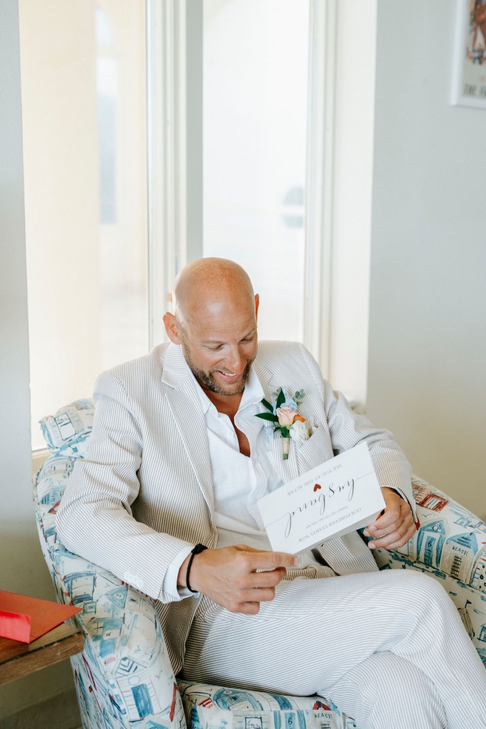 Smiling bald groom in linen suit reads a card while seated in a patterned chair near a bright window.