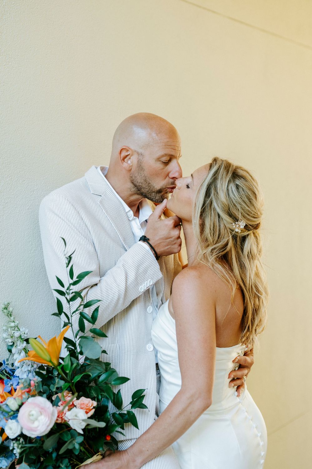 Bride and groom share a romantic kiss in wedding attire, holding a colorful floral bouquet against a neutral wall.