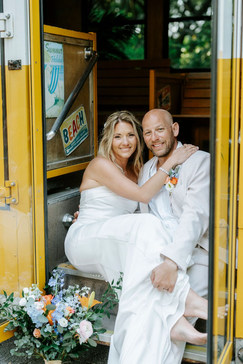 Happy bride and groom pose together in the doorway of a vintage yellow school bus on their wedding day.