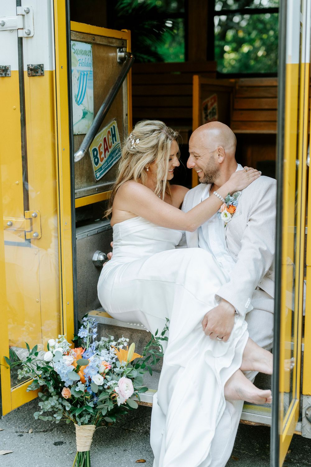 Bride and groom in white sitting in yellow bus doorway, sharing romantic moment with colorful wedding bouquet nearby.
