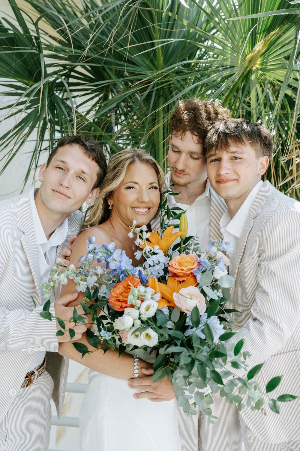 Bride holding colorful bouquet poses with three men in white suits near tropical palm trees at outdoor wedding.