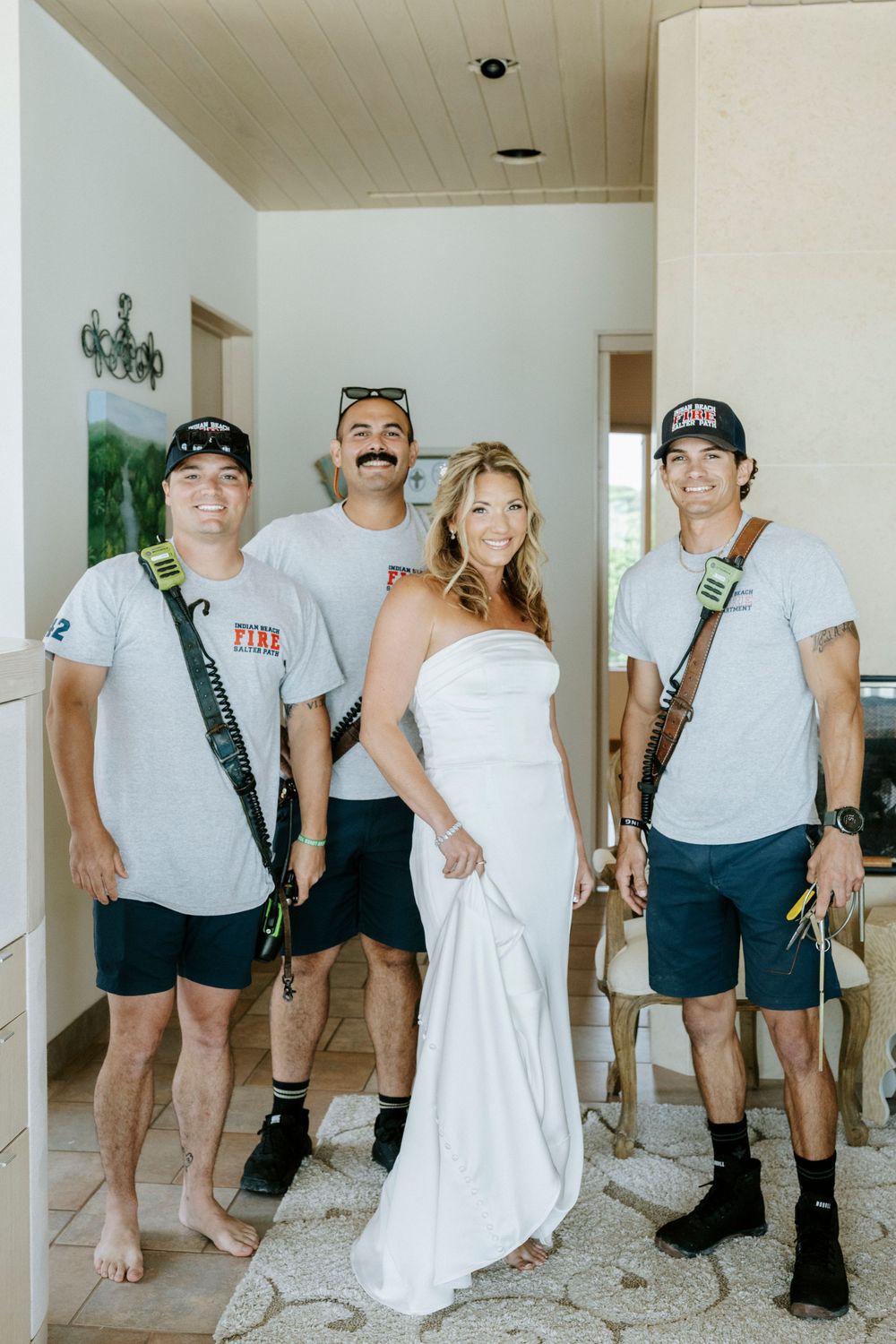 Bride in white strapless gown poses with three men in matching gray Fire department shirts indoors.