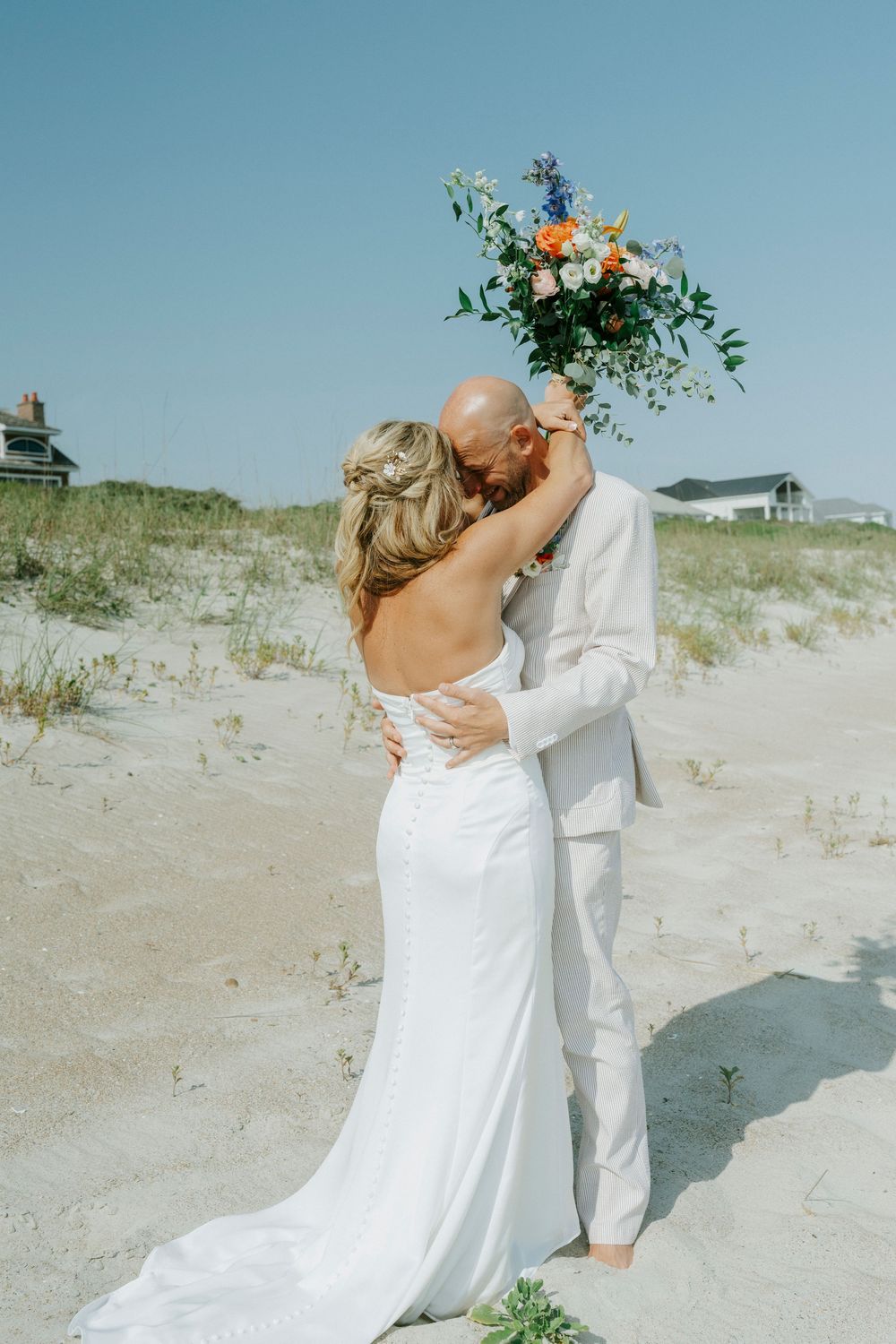 Bride and groom embracing on a sandy beach, she holds a colorful bouquet overhead, both dressed in white wedding attire.