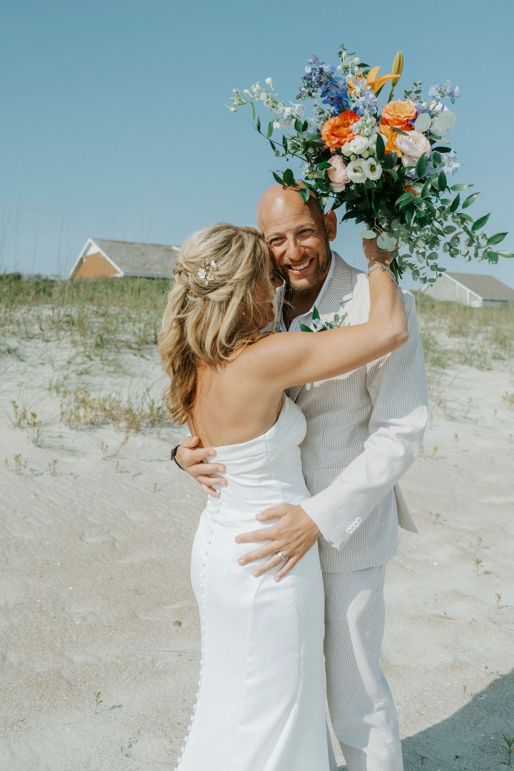 Smiling groom in white suit embraces bride on sandy beach, holding vibrant orange and blue floral bouquet.