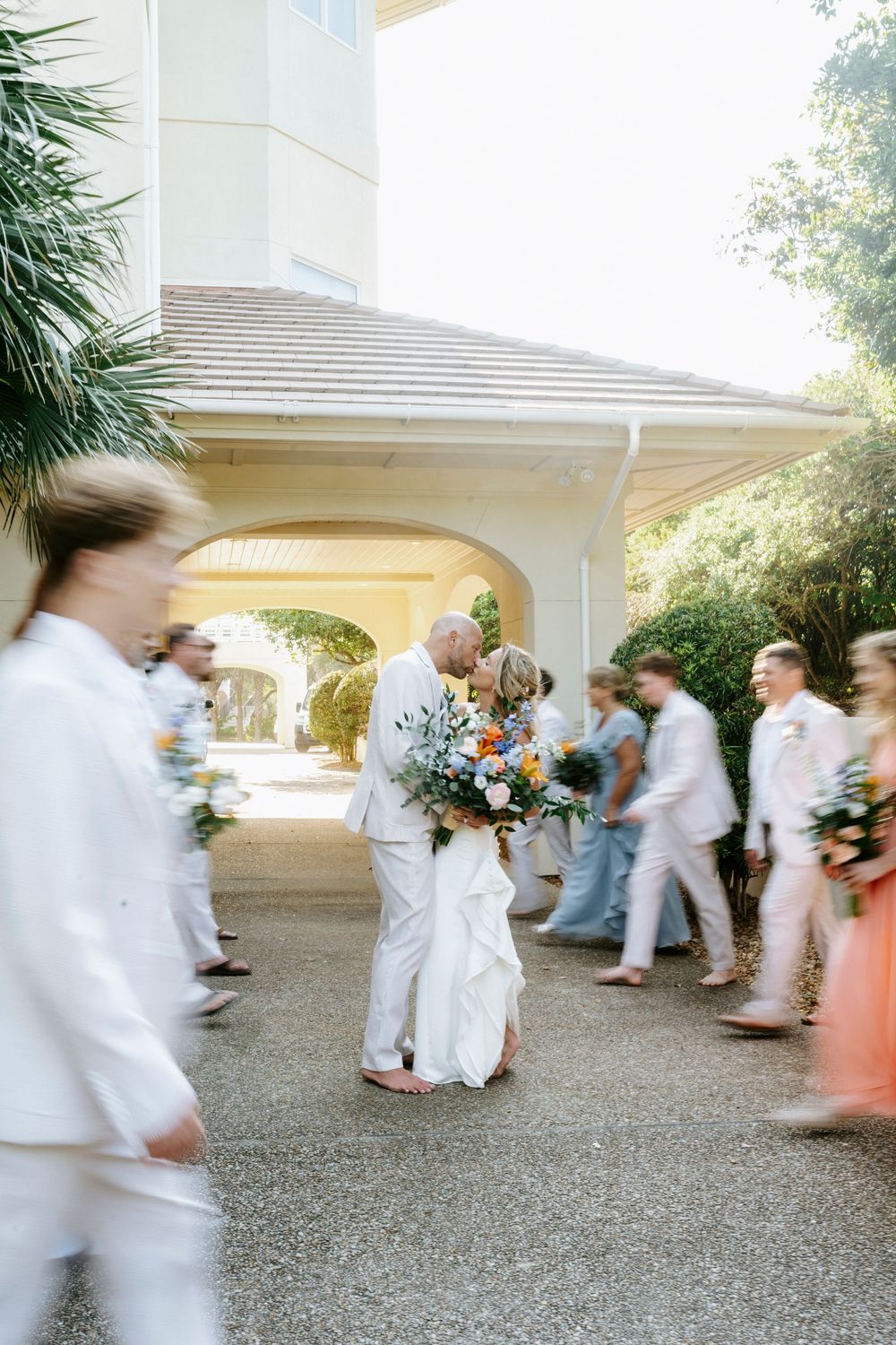 Bride and groom share a kiss surrounded by wedding party in motion blur at a sunny outdoor venue.