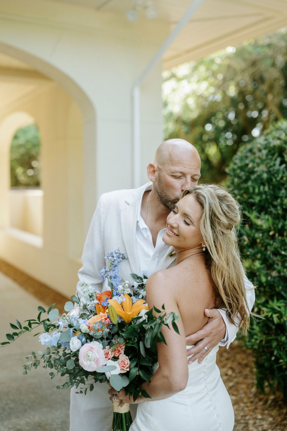 Bride and groom embrace outdoors, she holds a colorful bouquet of orange, blue, and pink flowers while he kisses her cheek.