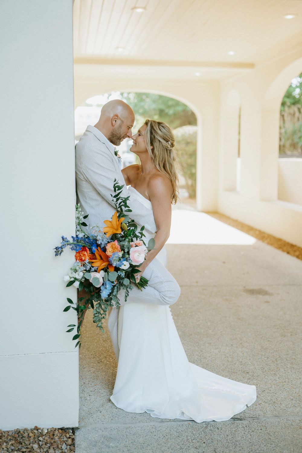 Bride and groom share romantic moment leaning against white pillar with colorful orange and blue bouquet.
