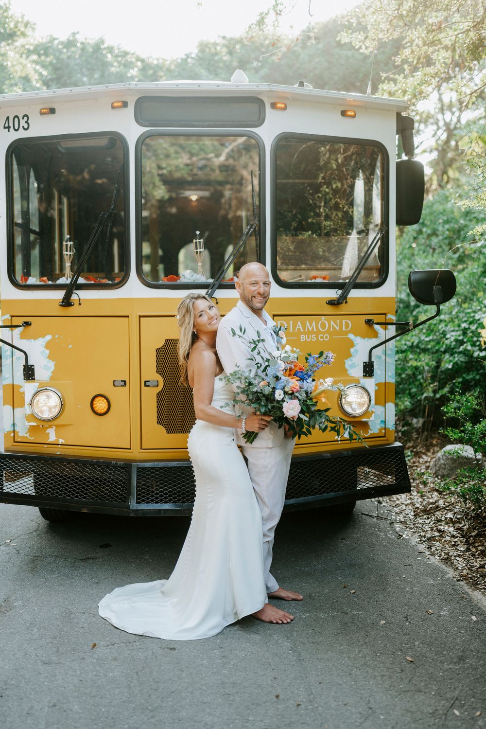 Bride and groom pose barefoot before a yellow Diamond Bus Co. trolley, bride holding a vibrant bouquet.