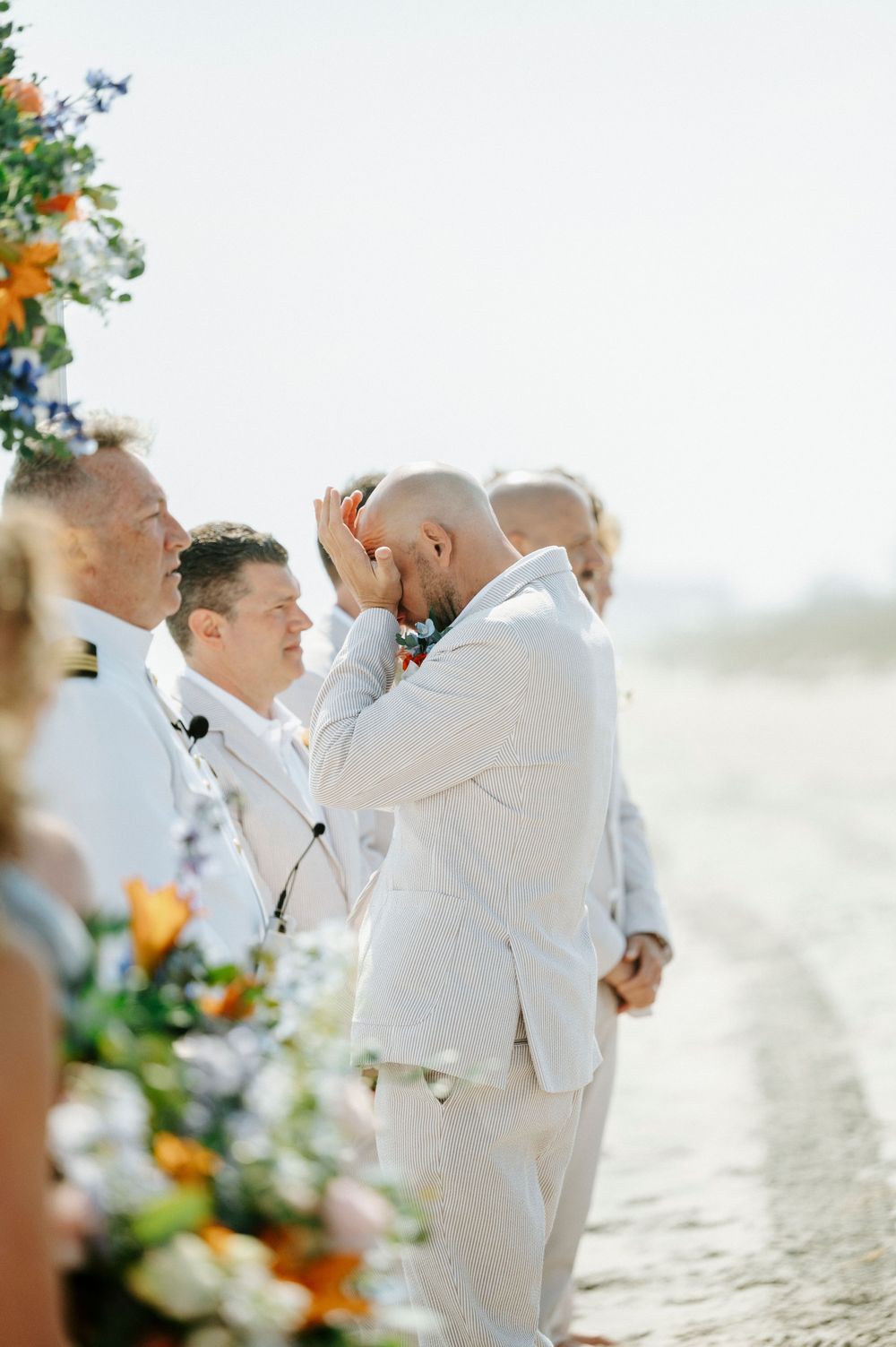 Emotional groom in white suit at beach wedding ceremony with colorful floral arrangements.