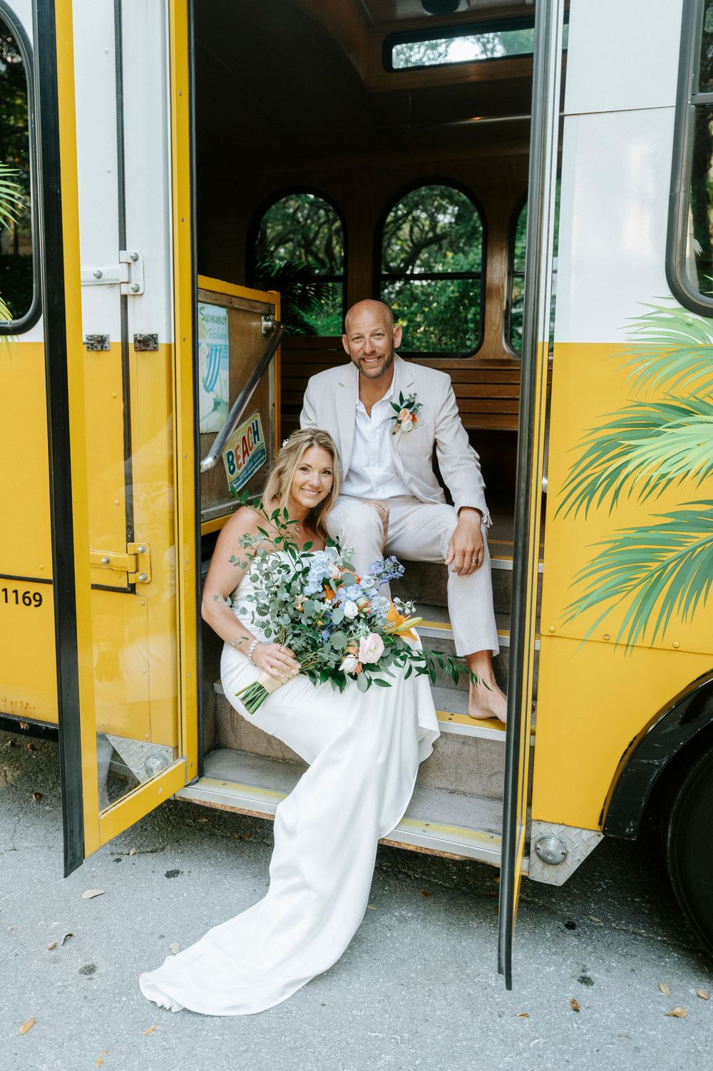Bride and groom posing on steps of yellow trolley, bride holding colorful bouquet.