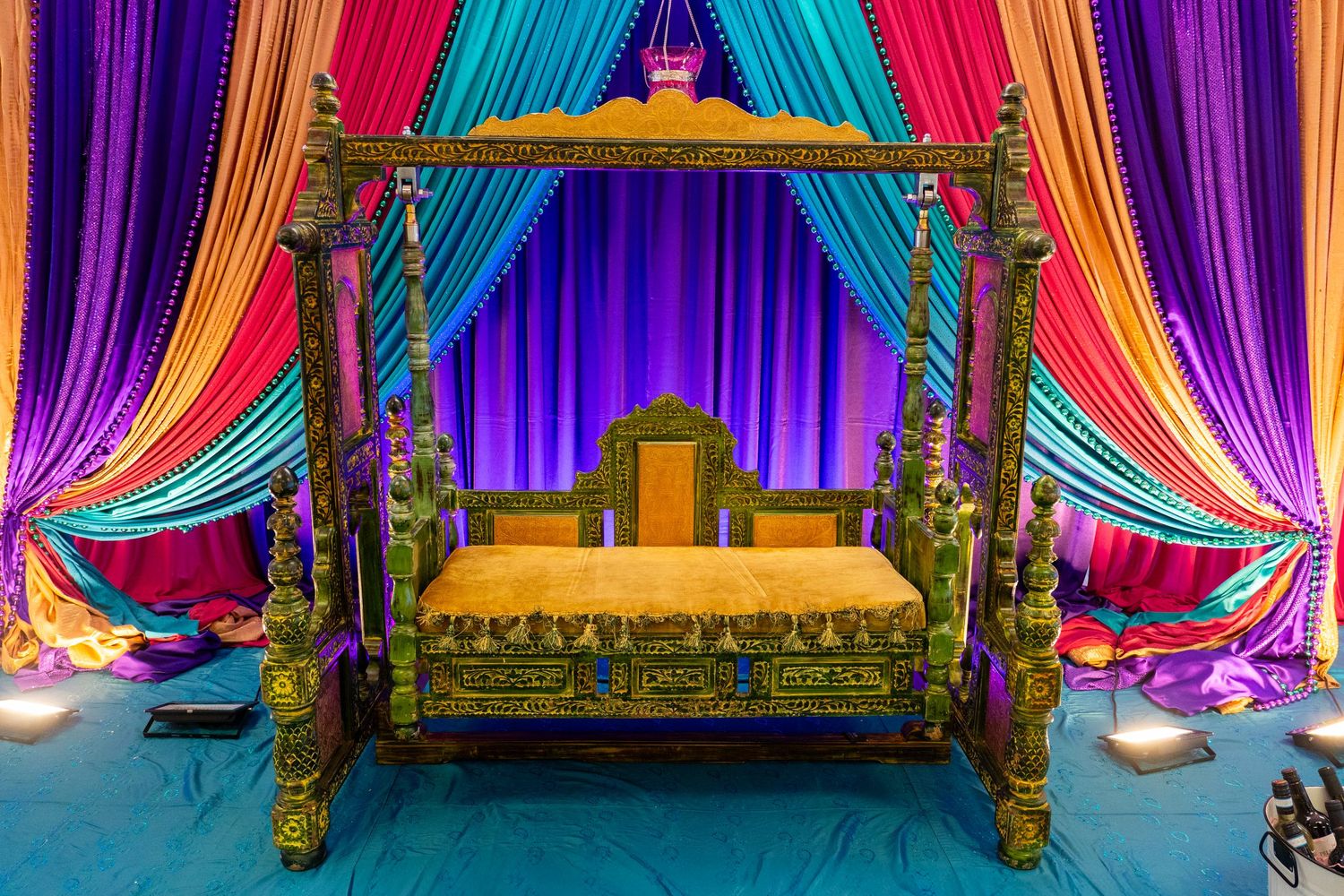 Ornate Indian wedding mandap with colorful silk drapes in purple, pink, and teal surrounding a decorated golden swing.