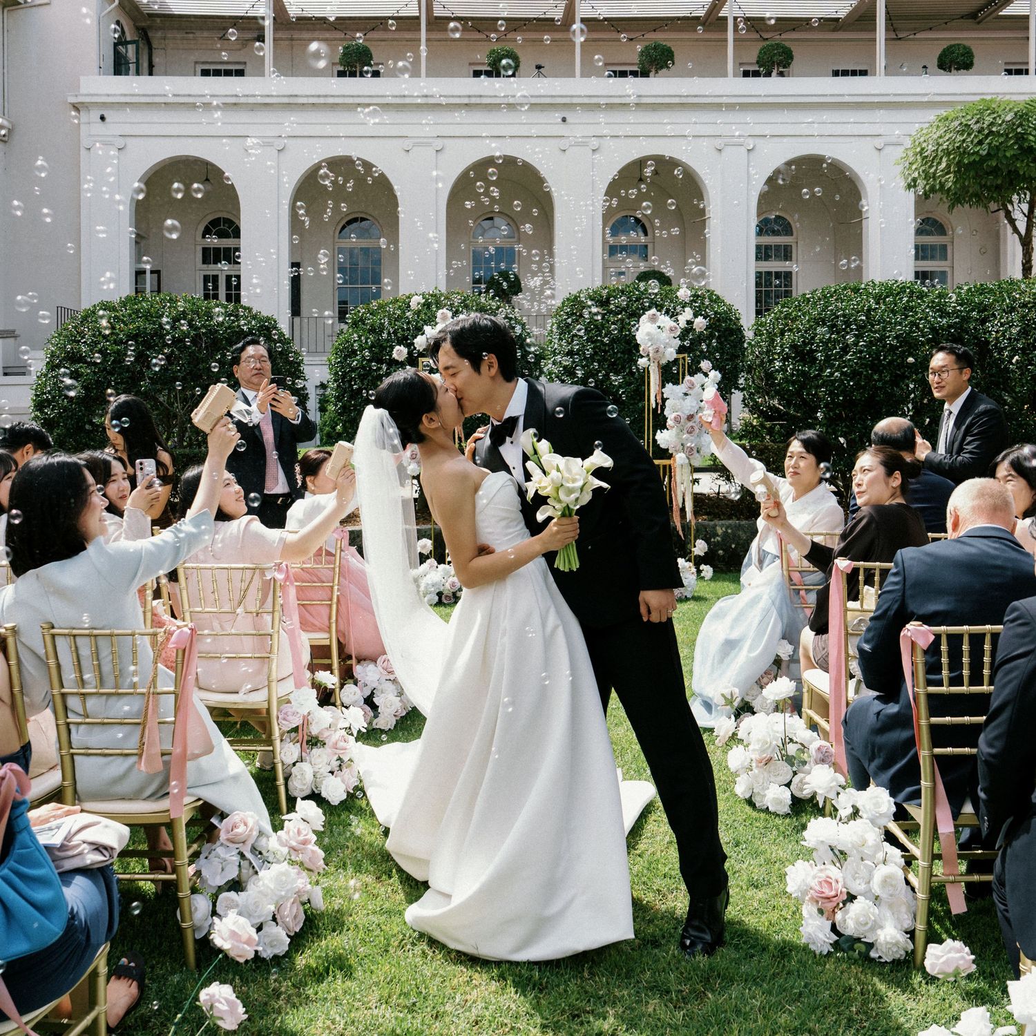 Bride and groom share a kiss at outdoor wedding ceremony with guests cheering and bubbles floating in elegant garden venue.