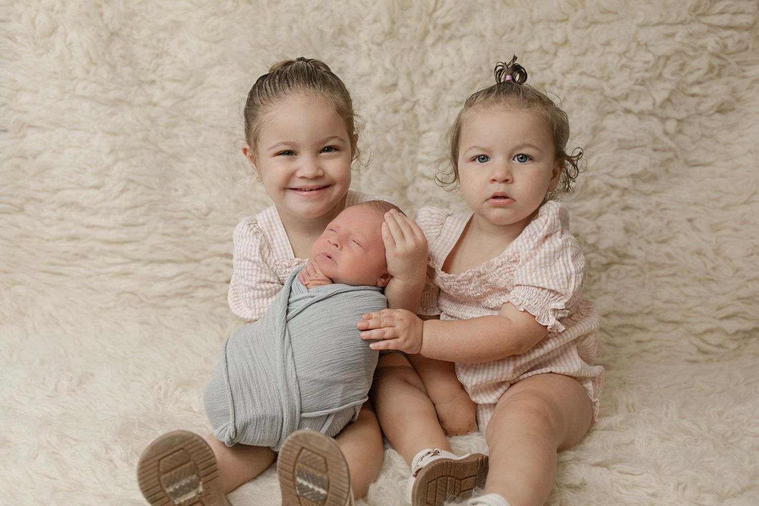 Two young sisters sit on fluffy rug gently holding their sleeping newborn baby sibling.