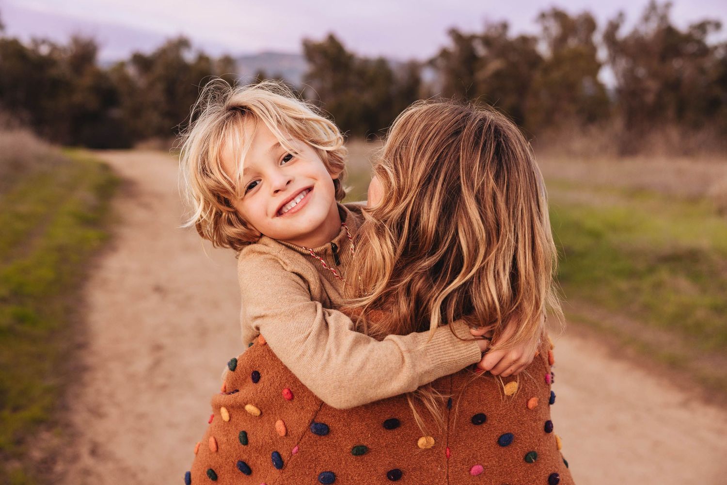Two young children hugging and smiling on a dirt path outdoors, wearing warm earth-toned clothing.