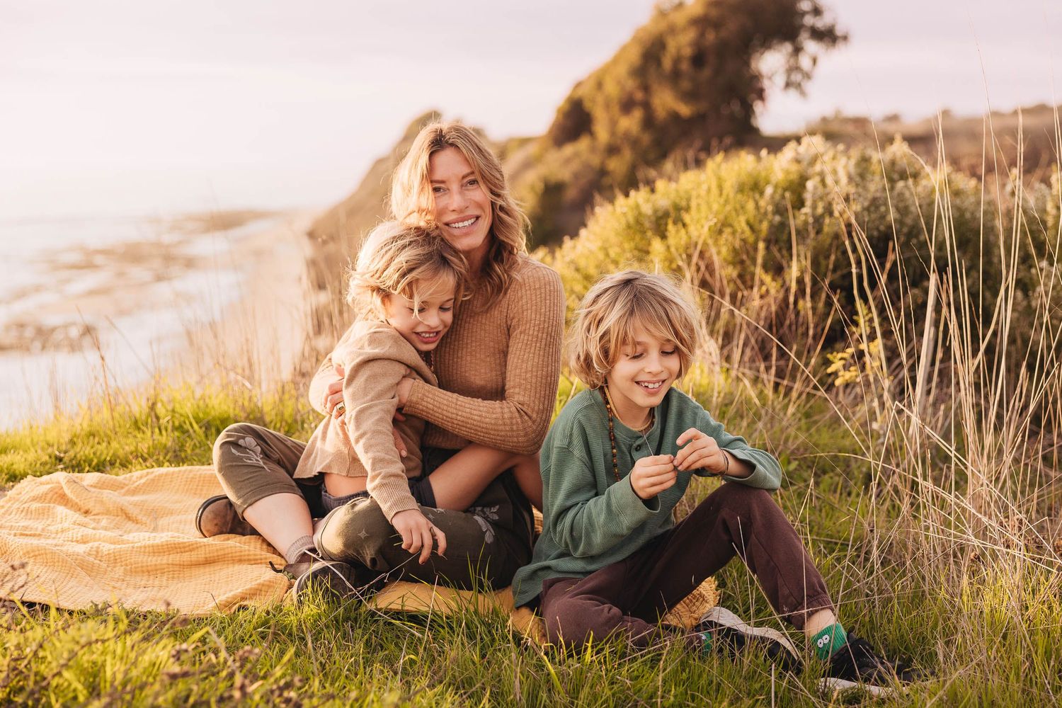 A smiling woman sits outdoors on a blanket with two children in a grassy coastal setting during golden hour.