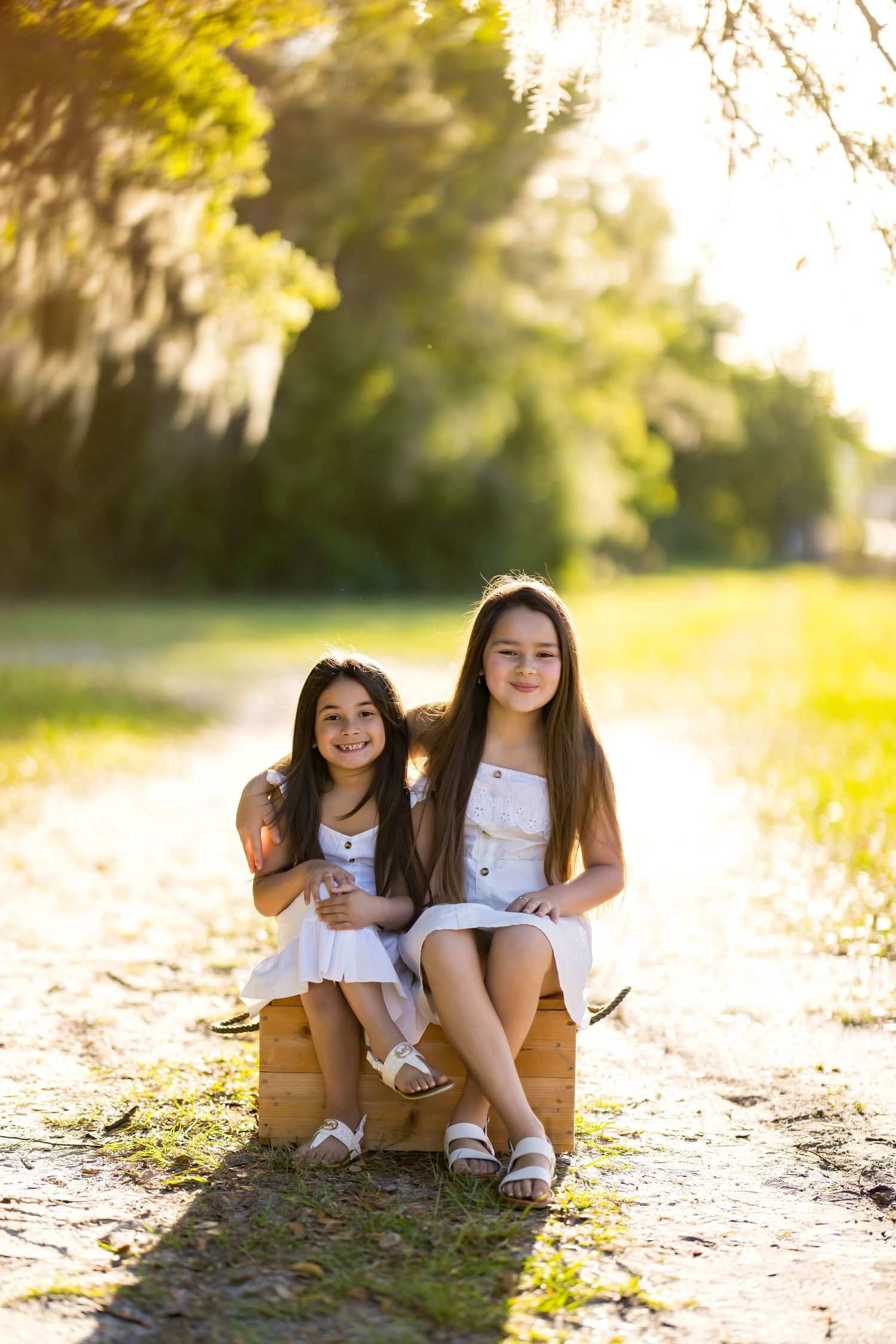 Two young girls in white dresses sit together on a wooden crate along a sunlit tree-lined path.