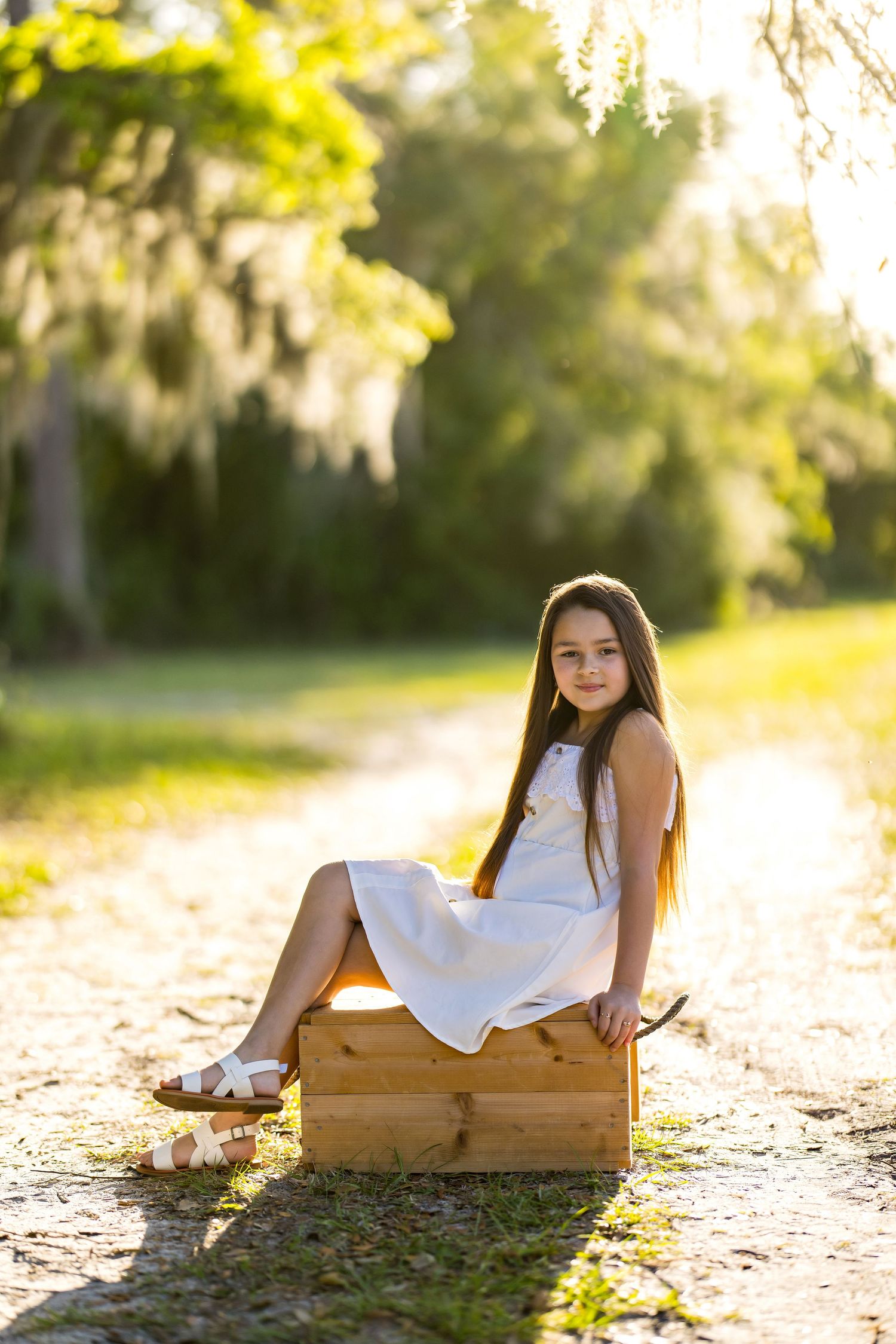 Young girl in white dress sitting on wooden crate along sunlit tree-lined path, golden hour outdoor portrait photography.