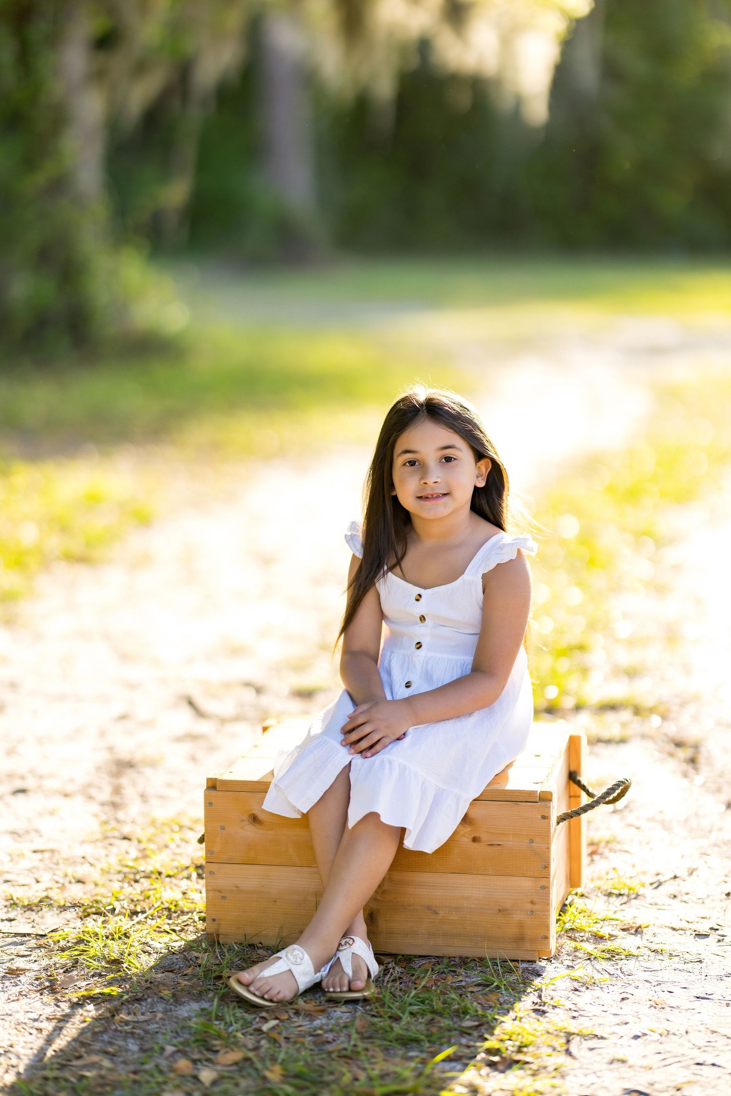 Young girl in white dress sitting on wooden crate outdoors on sunny path surrounded by lush green trees.