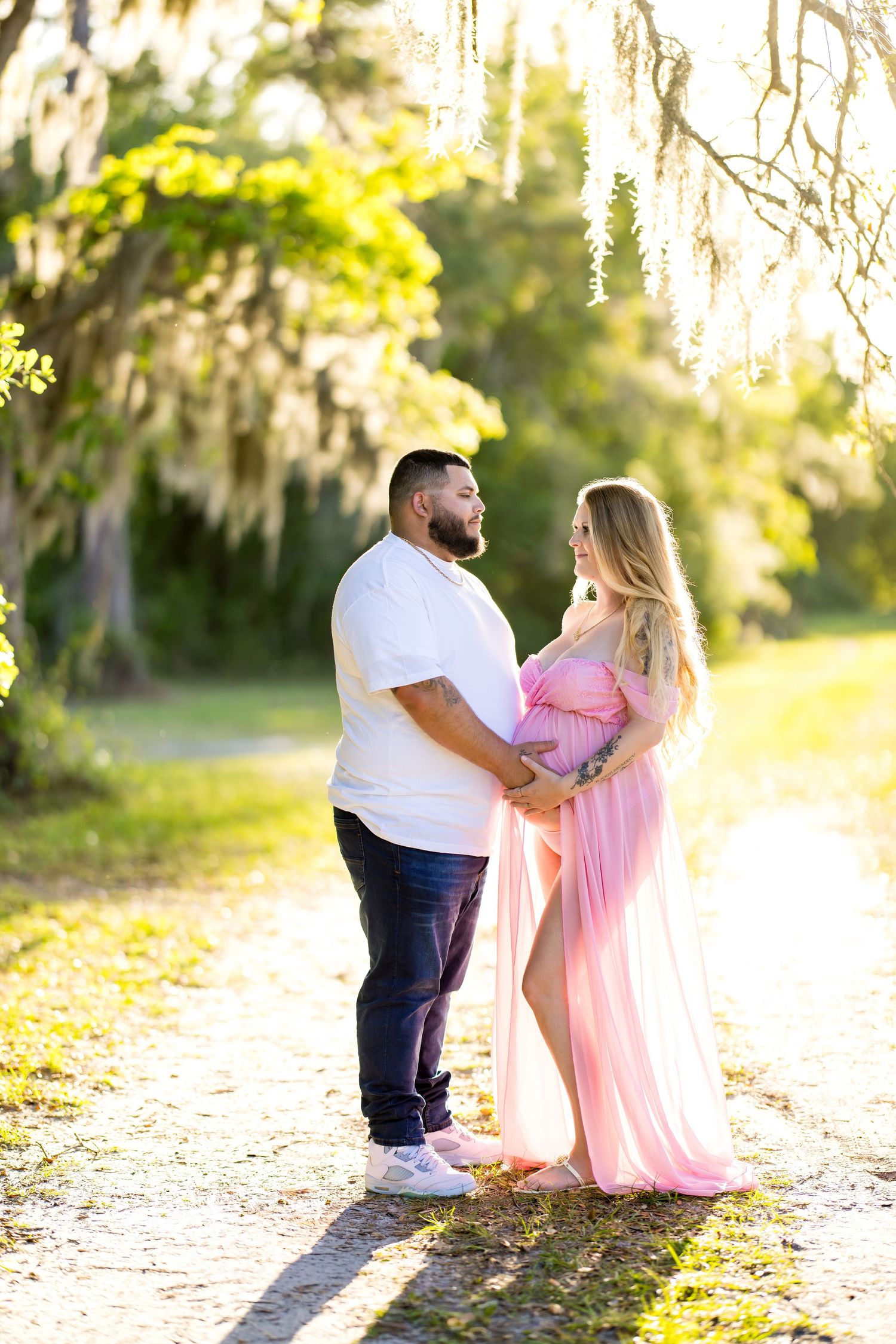 Couple holding hands outdoors during maternity photo shoot, woman in pink flowing gown, surrounded by moss-draped trees in golden light.