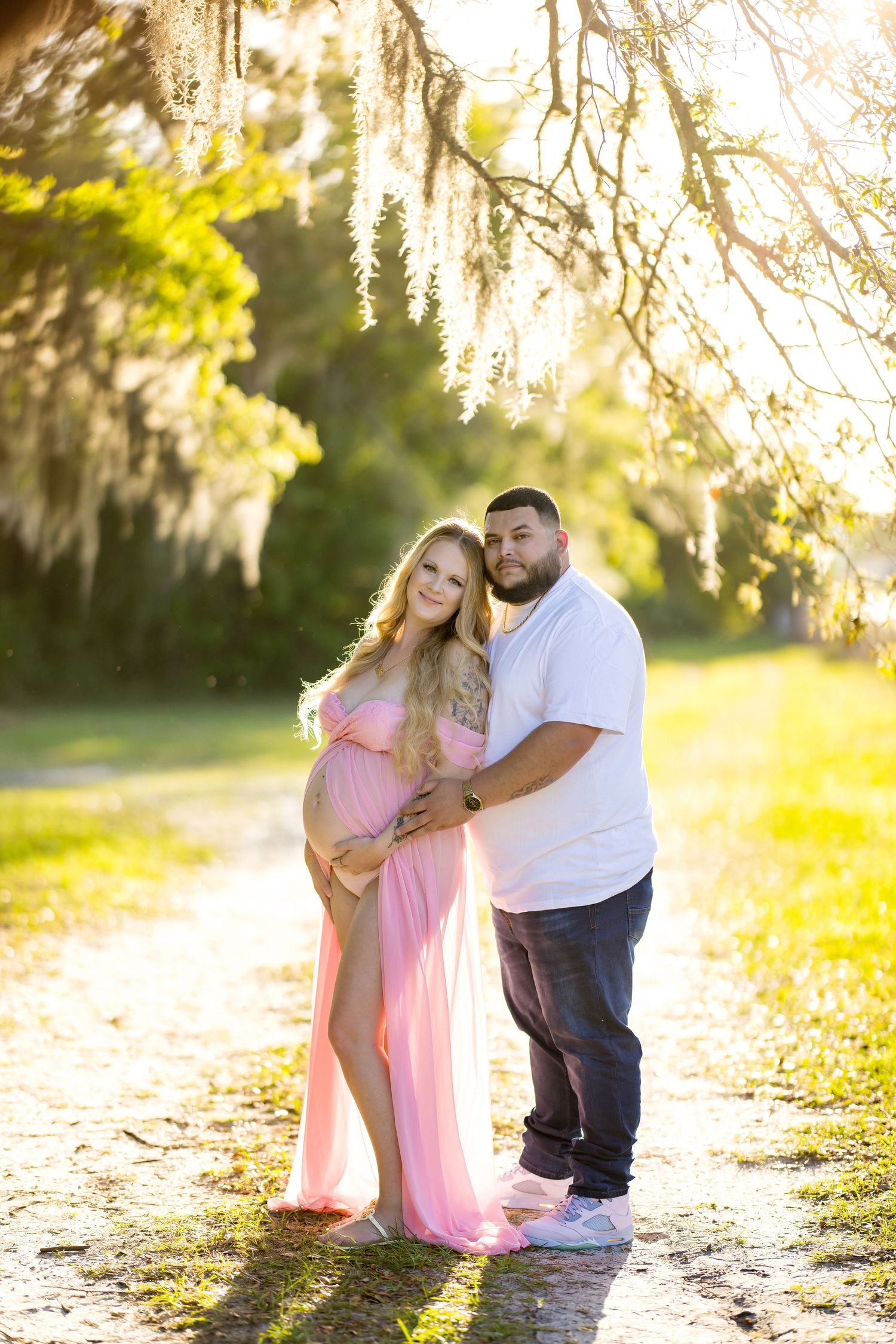 Couple poses outdoors for maternity photo, pregnant woman in pink gown, man in white shirt, sunlit trees with Spanish moss.