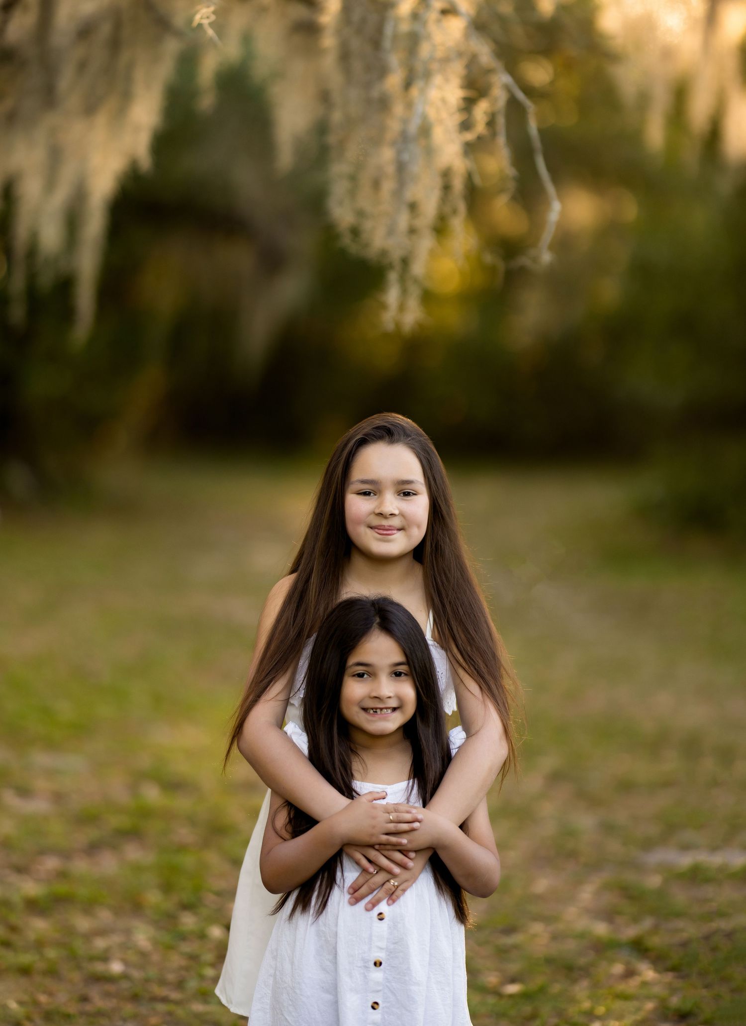 Two young sisters in white dresses embrace outdoors under Spanish moss-draped trees in a warm golden light.