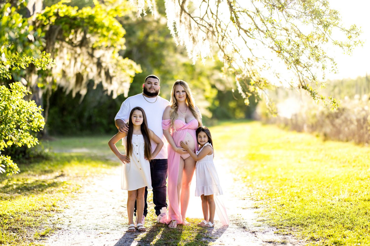 Pregnant woman poses with husband and two daughters on a sunny tree-lined path for a family maternity photo.