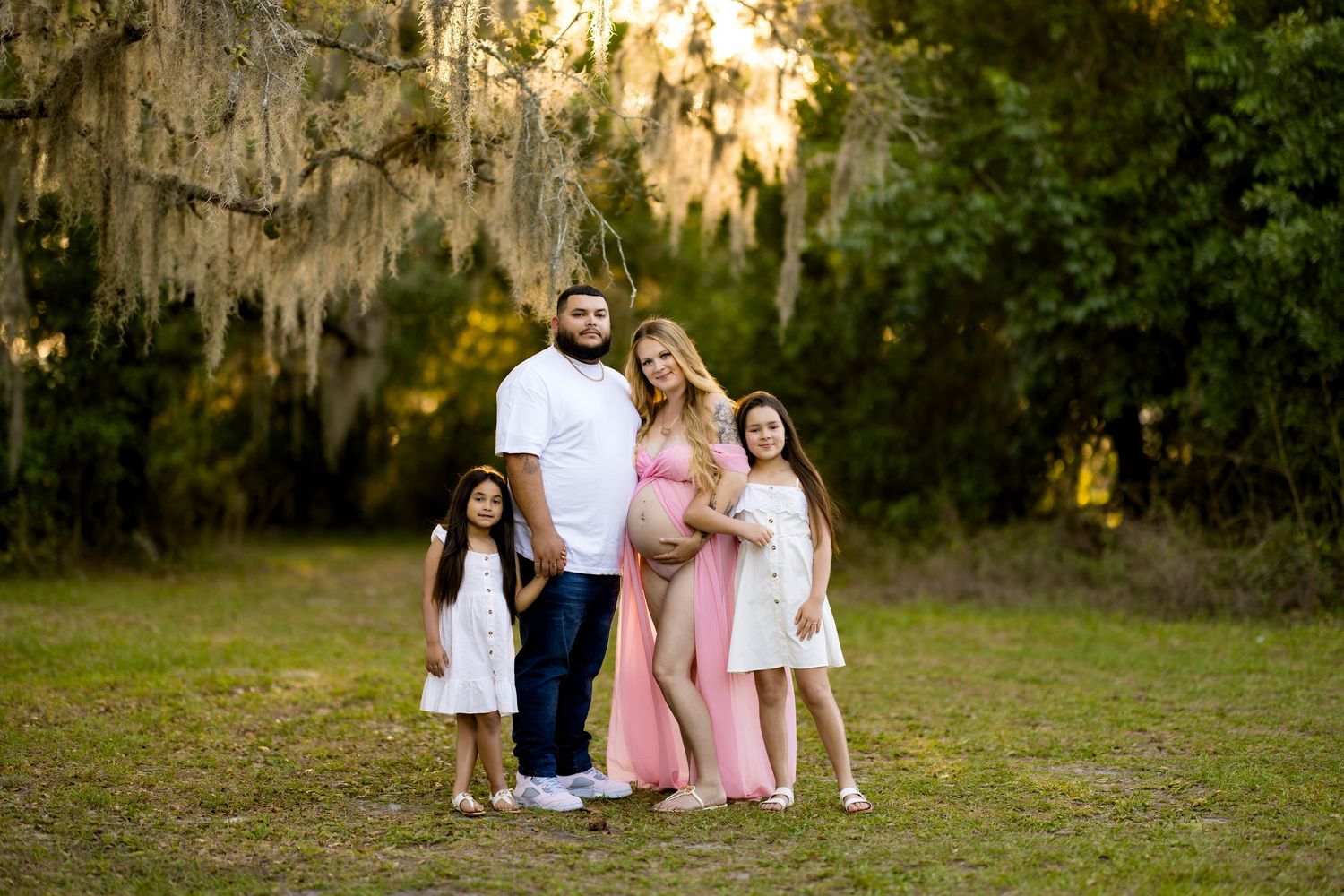 Pregnant woman in pink dress poses with family outdoors under moss-draped trees in a maternity photo session.