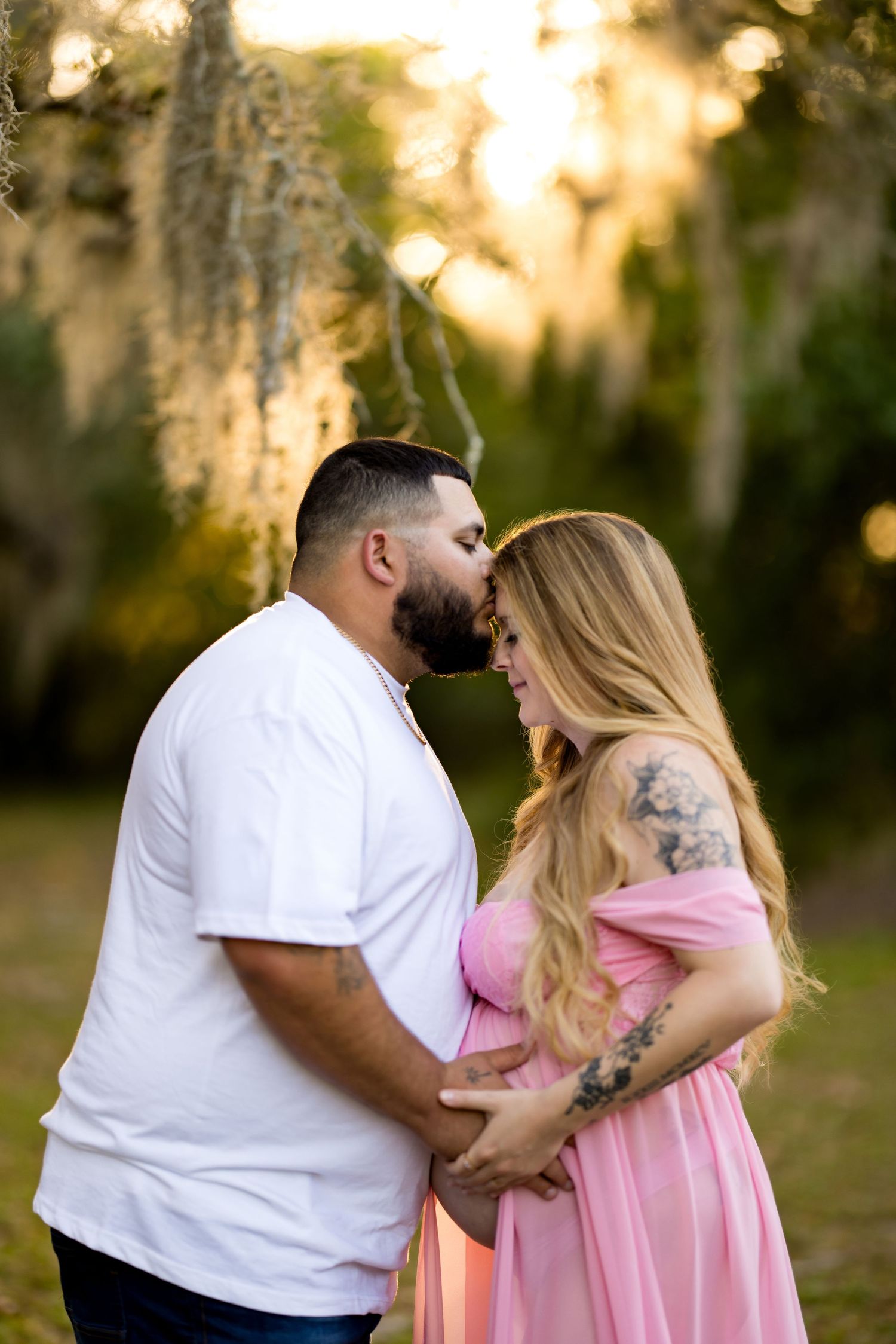 Couple sharing a tender kiss outdoors during a maternity photo session, woman in pink dress cradling baby bump at golden hour.