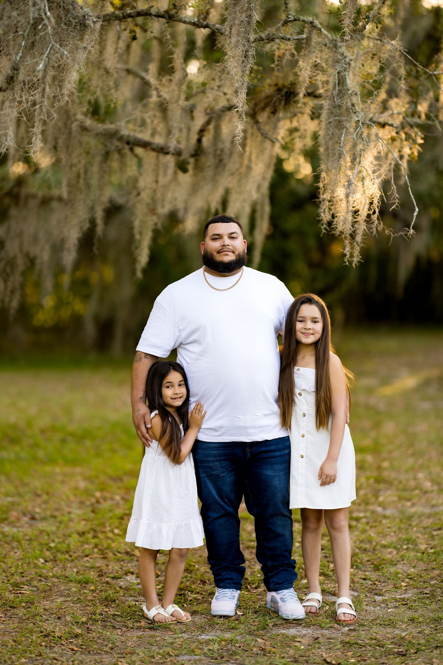 A man in a white shirt and jeans poses outdoors with two young girls in white dresses under Spanish moss-draped trees.