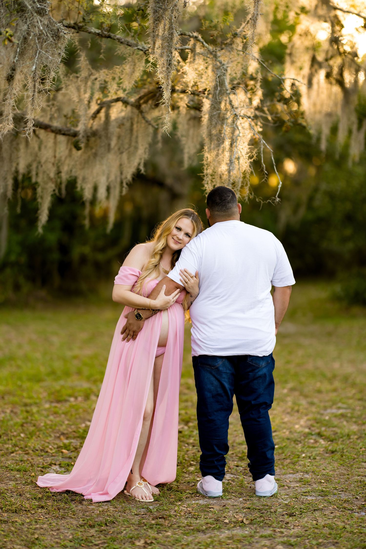 Pregnant woman in pink maternity gown with partner under Spanish moss trees during outdoor maternity photoshoot.