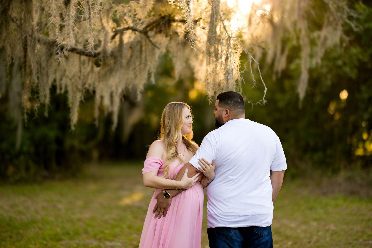 Couple standing together under Spanish moss-draped oak trees at golden hour, woman in pink maternity dress.