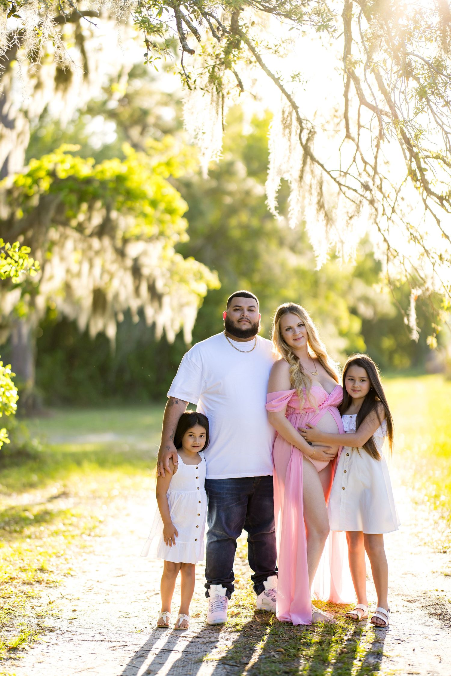 Pregnant woman in pink gown poses with family on sunlit path under Spanish moss-draped trees.