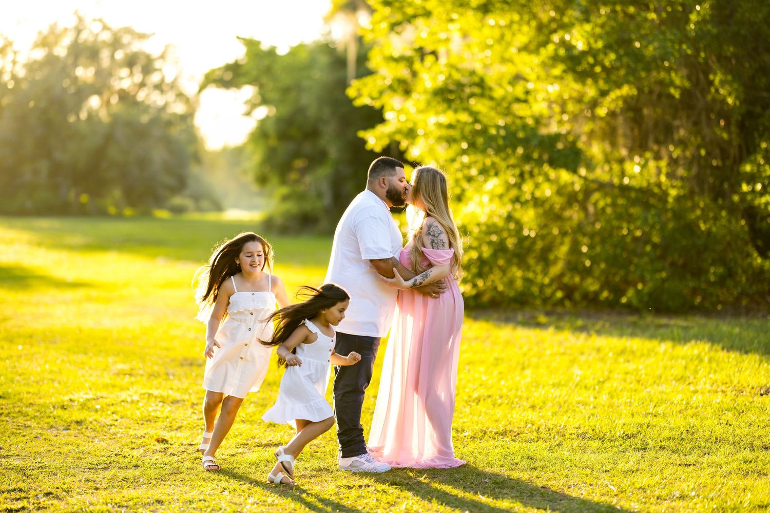 Family maternity photo session in a sunny green park, parents embracing while two young daughters run toward them.