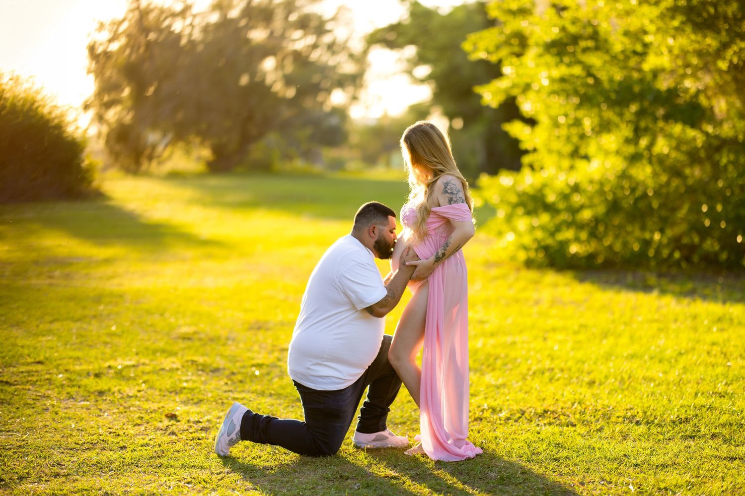 Man kneeling before pregnant woman in pink dress during golden hour maternity photoshoot in lush green park.
