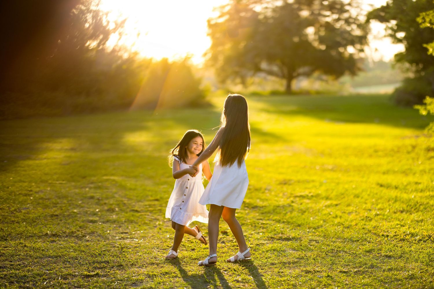 Two young girls in white dresses playing and spinning together in a sunny green park at golden hour.
