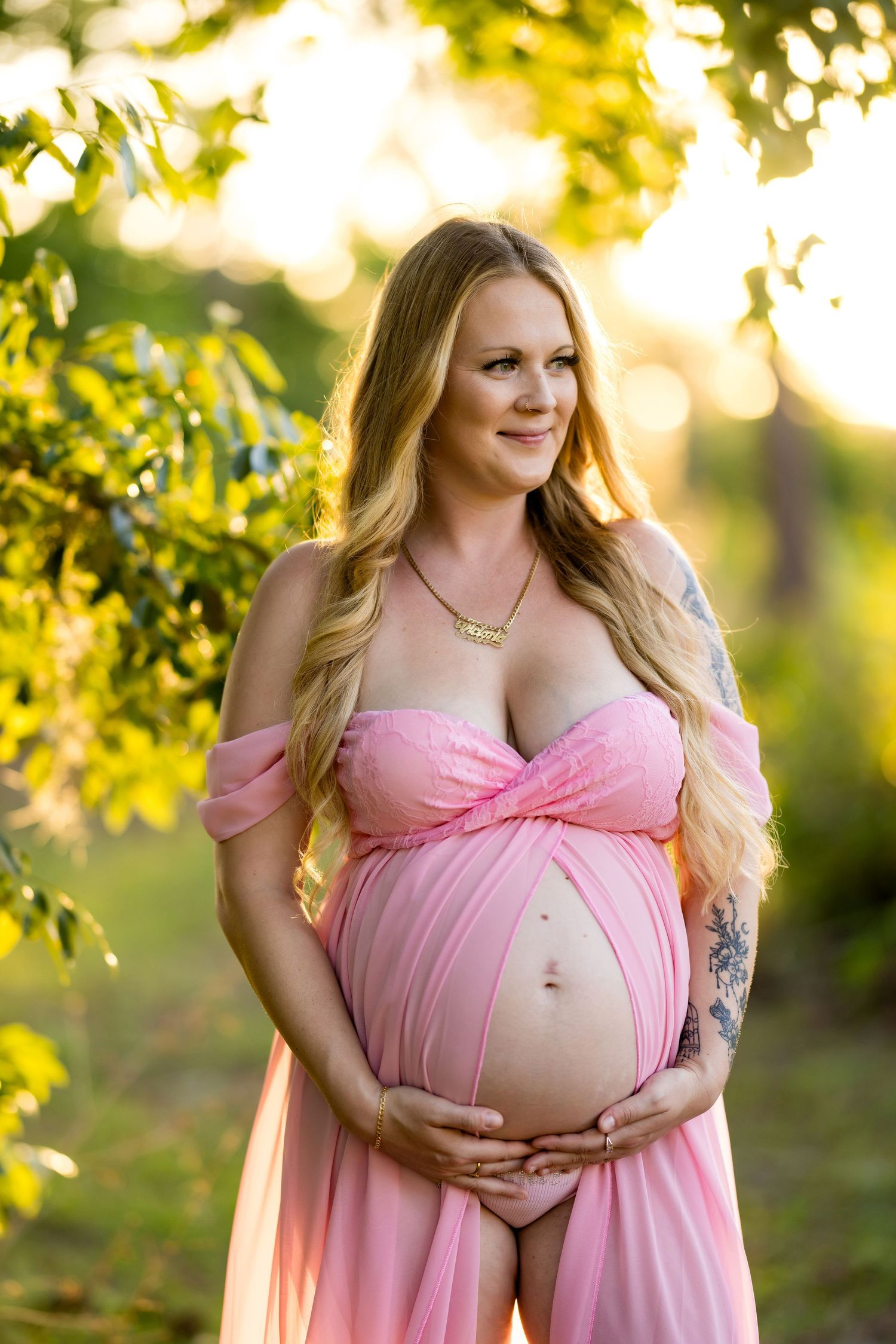 Pregnant woman in pink off-shoulder gown cradling baby bump outdoors in golden sunlight among green trees.