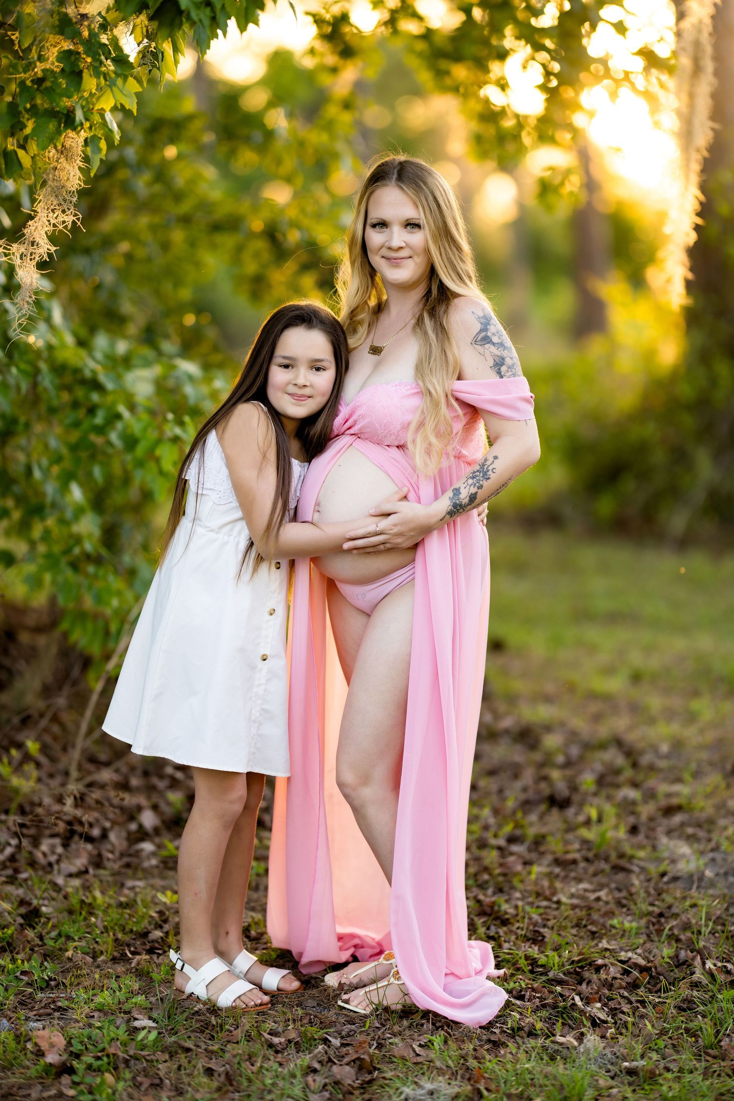 Pregnant woman in pink maternity gown poses outdoors with young girl in white dress at golden hour.