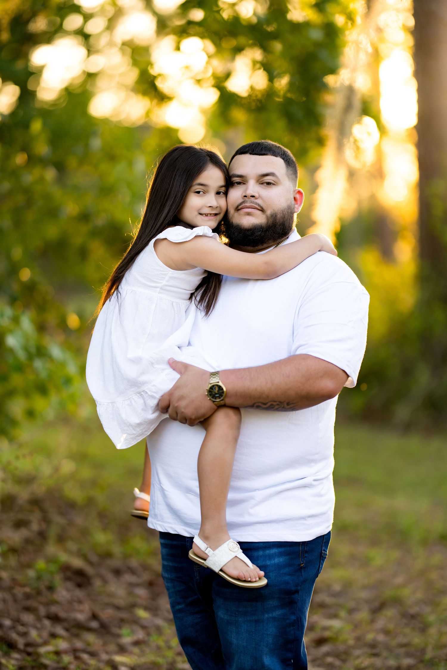 A father holds his young daughter in a warm outdoor portrait, both wearing white, surrounded by golden sunlit trees.