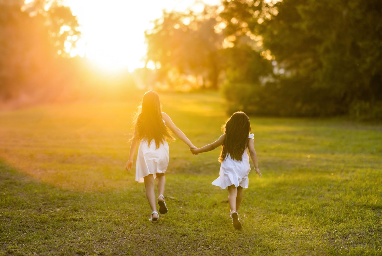 Two young girls in white dresses hold hands walking through a sunlit green field at golden hour sunset.