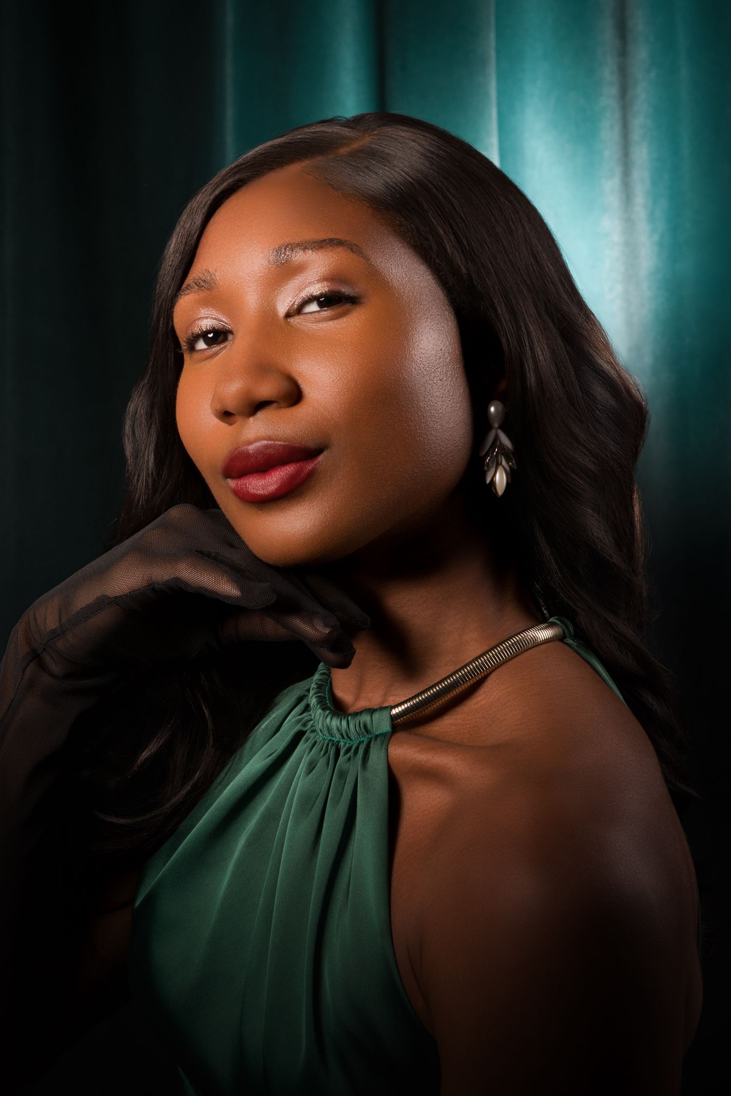 Elegant Black woman in green halter dress with gold necklace and leaf earrings poses against teal curtain backdrop.